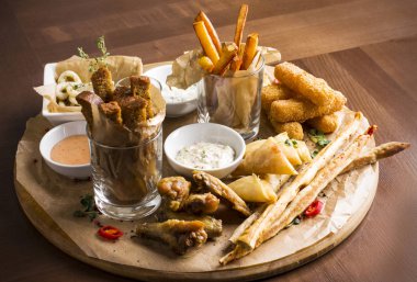 Farklı bira aperatifler: kalamar halkaları, patates kızartması, sıcak kanatları, ekmek ve ahşap tahta üzerinde soslar