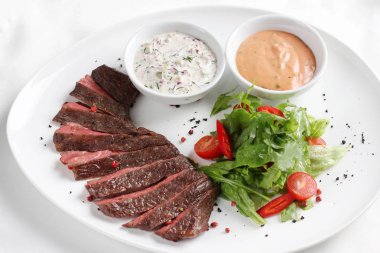 Dana biftek, orta pişmiş, taze sebzeler, otlar ve beyaz tabakta sos ile