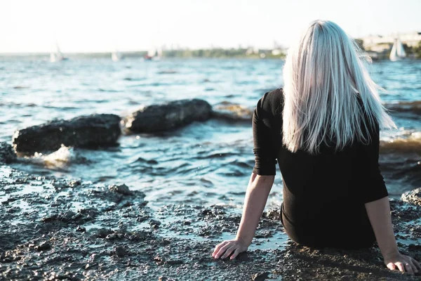 Het meisje met blond haar zit terug op de oever van lake zee en kijkt naar de boten — Stockfoto