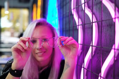 Oturan güzel sarışın kız neon duvarının yakınında çubuk ve gözlük tutar
