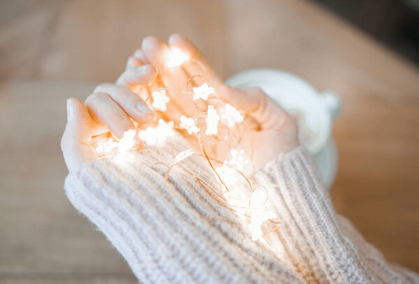 Молодая женщина в уютном бежевом свитере с гирляндами в руках за кофе и круассаном
