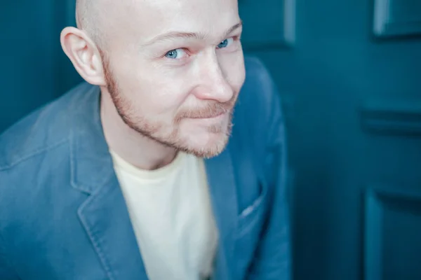 Attractive adult successful bald bearded man in suit looks into camera and smiles slyly on blue wall background
