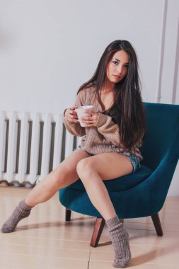 Samimi sıcak bej kazak holding mavi vintage sandalyede oturuyor çay fincan koyu uzun saçlı şehvetli gülümseyen Asyalı kız genç kadın portresi