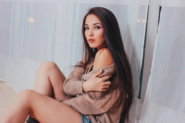 Candid portrait of sensual smiling asian girl young woman with dark long hair in cozy beige sweater on white curtain background