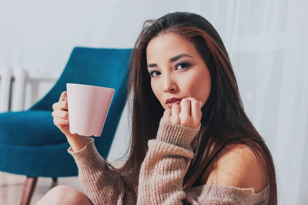 Candid portrait of sensual smiling asian girl young woman with dark long hair in cozy beige sweater holding cup of tea on white curtain background