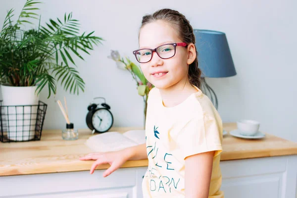 Pretty smiling little girl in glasses and pigtails sitting with book at the table in her room — Stock Photo, Image