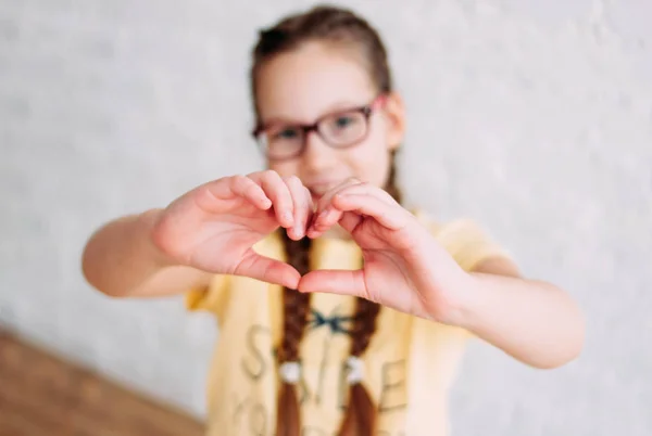 Happy tweens girl in yellow t-shirt making heart by hands on the white wall background, selective focus