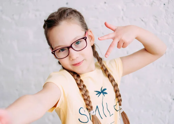 Cute tweens girl in yellow t-shirt with funny pigtails taking selfie on frontal camera on the white brick wall background