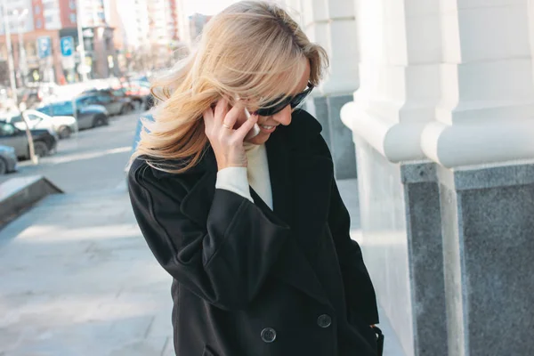 Mulher loira elegante vestindo casaco preto e óculos de sol falando no telefone celular na rua — Fotografia de Stock