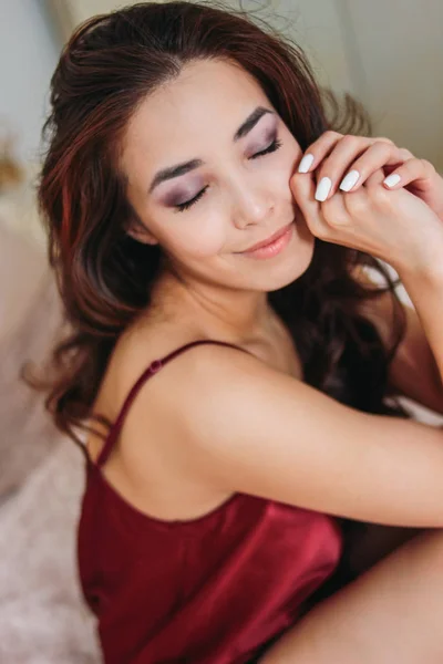 행복 한 관능적인 웃는 소녀 아시아 젊은 여 자가 어두운 긴 곱슬 머리에 빨간 속옷에 앉아 침대에 — 스톡 사진