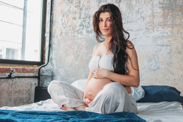 Красивая беременная девушка молодая женщина с длинными вьющимися волосами сидит в позе лотоса на кровати. Беременность йога и природа этническая красота концепции — стоковое фото