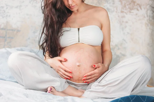 Oříznout fotografii krásné těhotné dívky s dlouhými kudrnatými vlasy, které sedí v lotosové pozici na posteli. Těhotenský józ a přírodní koncepce krásy — Stock fotografie