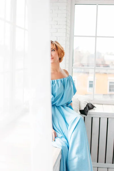 Young charming smiling blonde short hair woman in blue summer long dress sitting on window sill — ストック写真