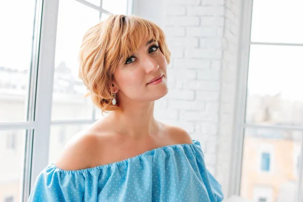 Junge charmante lächelnde blonde Frau mit kurzen Haaren in blauem Sommerkleid in der Nähe des Fensters im weißen Zimmer — Stockfoto