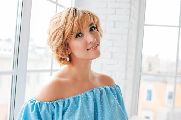 Young charming smiling blonde short hair woman in blue summer dress near window in white room — 스톡 사진