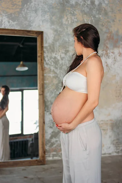 Vysoká krásná těhotná mladá žena s dlouhými kudrnatými vlasy, stojící před zrcadlem v podkrovním studiu — Stock fotografie