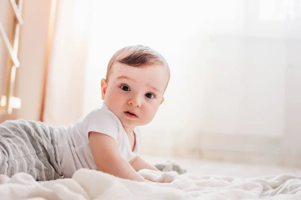 Обаятельный маленький мальчик 6 месяцев смотрит в камеру на белом одеяле в доме — стоковое фото