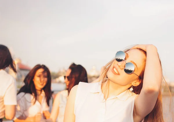 Positieve mooie gelukkig rood haired meisje in de spiegel zonnebril met vrienden op blauwe hemel achtergrond, zomer zonsondergang tijd — Stockfoto