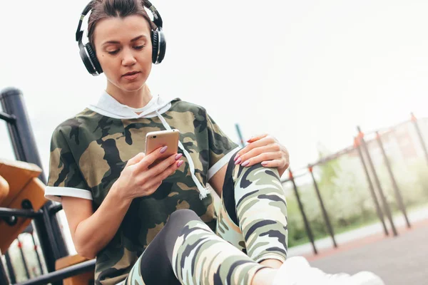 Attrayant jeune femme en forme dans les écouteurs et les vêtements de sport de couleur militaire faire de l'entraînement avec mobile sur la zone d'entraînement de rue. Le mode de vie sain dans le concept de ville — Photo