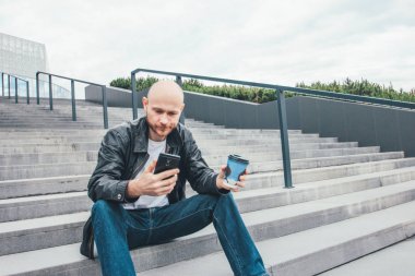 Yetişkin kel kasvetli adam kağıt fincan kahve içme ve şehir sokakta merdivenlerde oturan cep telefonu kullanarak
