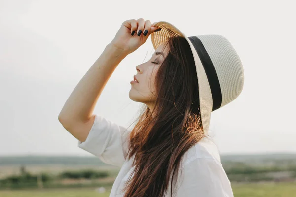 Portret van charmante zorgeloze lang haar Aziatisch meisje in witte kleren en stro hoed geniet van het leven in het veld bij zonsondergang. Gevoeligheid voor natuur concept — Stockfoto