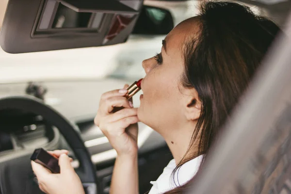 Mooie succesvolle elegante brunette jonge vrouw die haar lippen met rode lippenstift in de auto schilderij — Stockfoto