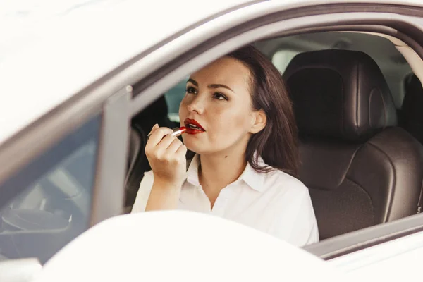 Mooie succesvolle elegante brunette jonge vrouw die haar lippen met rode lippenstift in de auto schilderij — Stockfoto