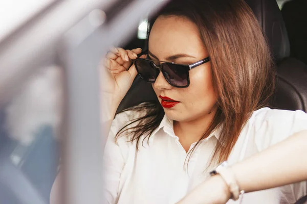 Mooie succesvolle elegante brunette jonge vrouw met rode lippen in zonnebril rijden personenauto — Stockfoto