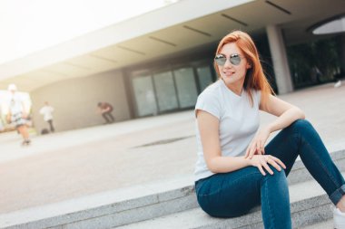 Şehirde sokakta oturan rahat giysiler içinde yuvarlak güneş gözlüğü çekici kızıl saçlı gülümseyen kız