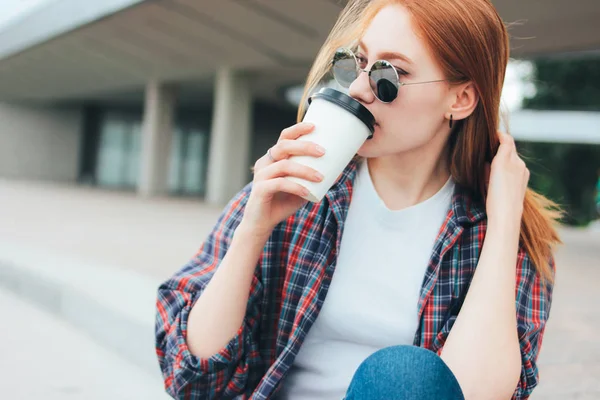 Attractive redhead smiling girl in round sunglasses casual wearing drinking coffee to go sitting on street in city — Stock Photo, Image