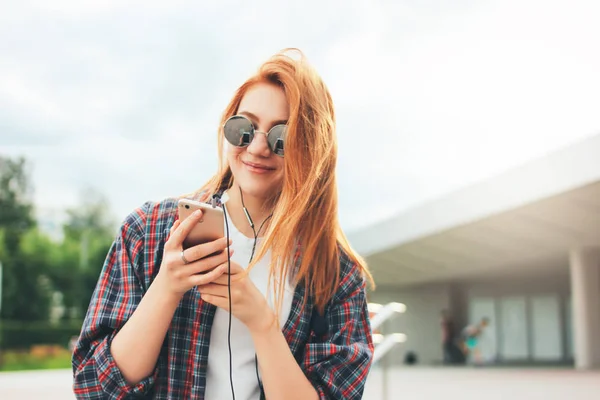 Привлекательная рыжая улыбающаяся девушка в круглых солнцезащитных очках с телефоном в руках в повседневной одежде слушает музыку на улице в городе — стоковое фото