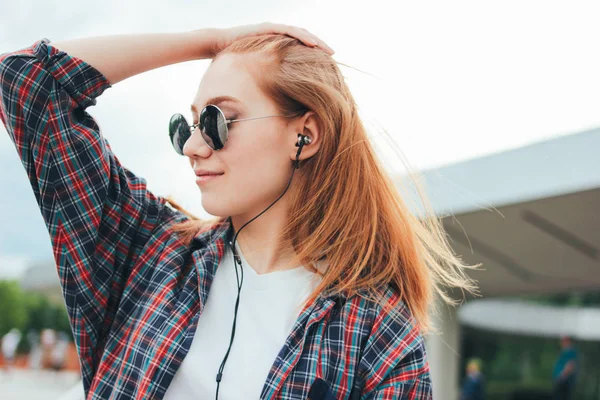 Jolie rousse souriante fille dans des lunettes de soleil rondes avec le téléphone dans ses mains dans des vêtements décontractés écouter de la musique dans la rue en ville — Photo