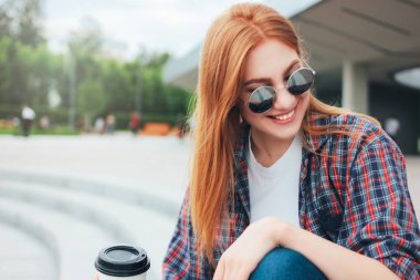Şehirde sokakta oturan rahat giysiler içinde ellere gitmek için kahve fincan ı ile yuvarlak güneş gözlüğü içinde Çekici kızıl saçlı gülümseyen kız