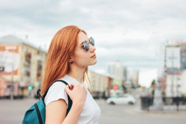 Şehirde sokakta sırt çantası ile rahat giysiler içinde güneş gözlüğü çekici kızıl gülümseyen kız