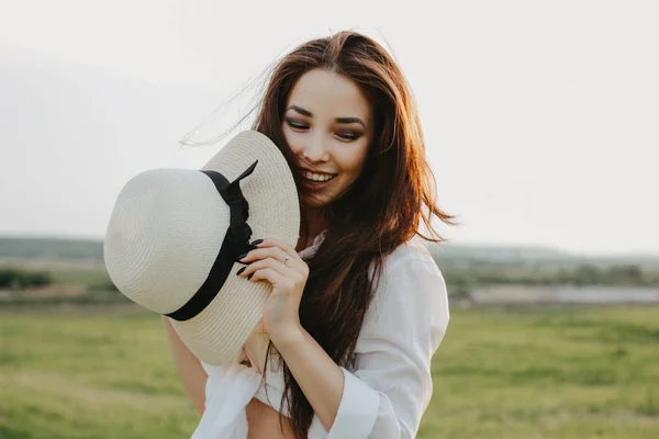 Portret van charmant zorgeloos lang haar Aziatische meisje in witte kleren en stro hoed geniet van het leven in het natuurgebied bij zonsondergang. Gevoeligheid voor natuur concept — Stockfoto