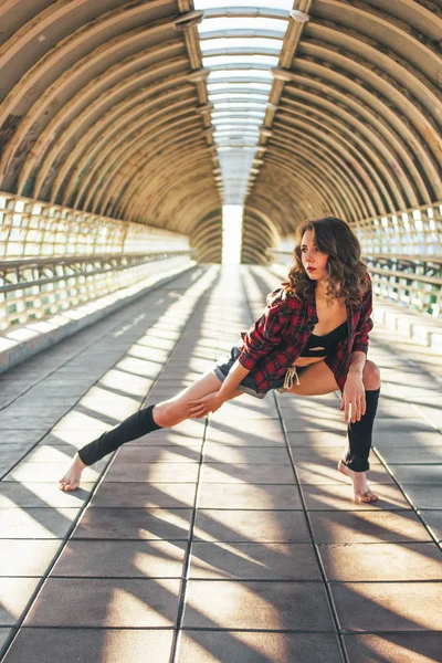 Sottile giovane bruna donna ballerina di strada ginnasta si siede in posa spacchi sul ponte di strada all'alba — Foto Stock