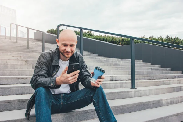 Adulto careca sorrindo homem bebendo café de copo de papel e usando telefone celular sentado em escadas na rua da cidade — Fotografia de Stock