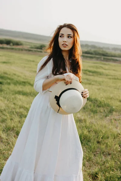 Bella spensierata capelli lunghi ragazza asiatica in vestiti bianchi e cappello di paglia gode la vita nel campo naturale al tramonto. Sensibilità al concetto di natura — Foto Stock