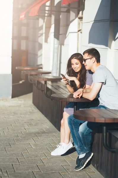 Jovem casal adolescentes amigos vestidos em estilo casual sentados juntos e usando móvel na rua da cidade — Fotografia de Stock