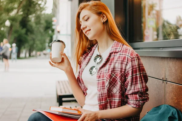 Привлекательная рыжая улыбчивая студентка оделась в повседневную одежду с чашкой кофе, чтобы пойти в руки на улице в городе — стоковое фото