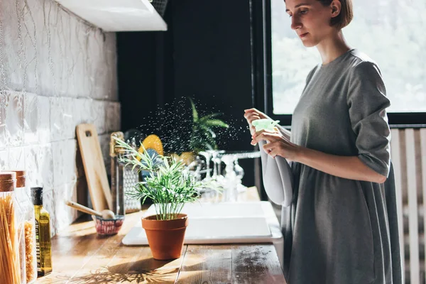 young woman sprays water on houseplant in the kitchen, slow life