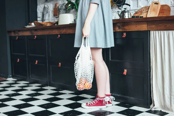 Молодая женщина в сером платье с вязаным тряпичным мешком мешок покупатель на кухне, ноль отходов, медленный срок службы — стоковое фото