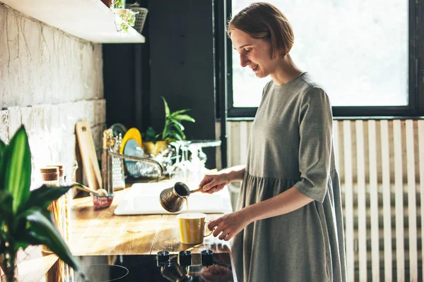 Jovem mulher em vestido cinza derrama café fresco natural em uma caneca na cozinha, de manhã, luz dura — Fotografia de Stock