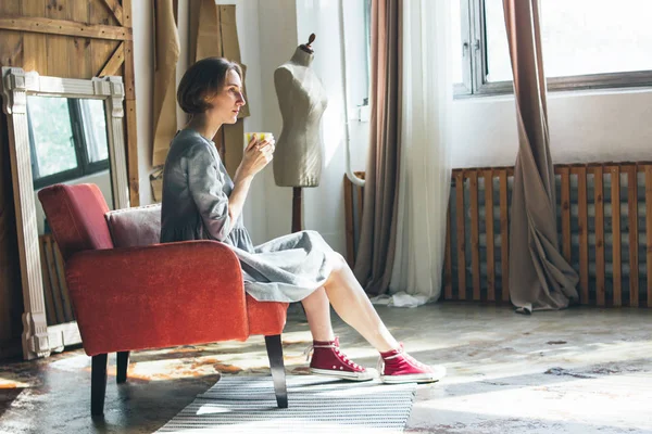 Giovane donna in semplice abito grigio con coppa gialla seduta su sedia in studio d'arte, ritratto a figura intera — Foto Stock