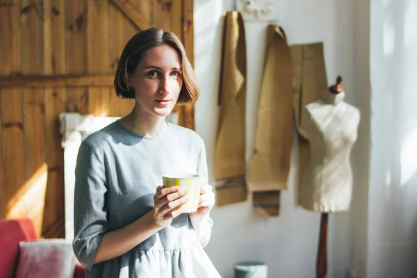 Jovem mulher em vestido cinza simples com copo amarelo sentado na cadeira em estúdio de arte, retrato de comprimento total — Fotografia de Stock