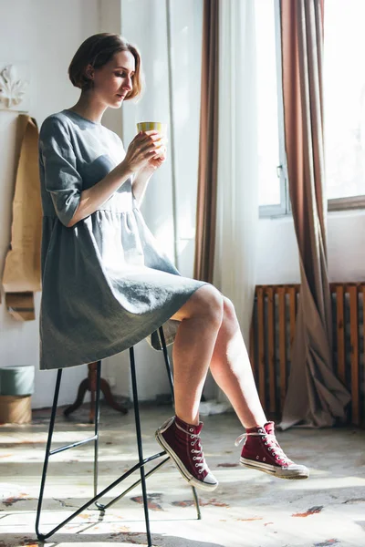 Jovem mulher em vestido cinza simples com copo amarelo sentado na cadeira em estúdio de arte, retrato de comprimento total — Fotografia de Stock