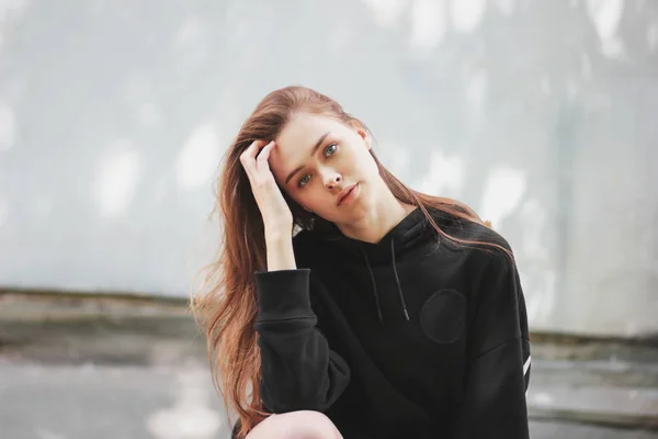 Candid portret van jonge mooie lange haren meisje fashion model hipster in zwarte hoodie op muur achtergrond — Stockfoto