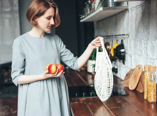 Young woman in grey dress pulls apples out of knitted rag bag string bag shopper in the kitchen, zero waste, slow life — Stock Photo, Image