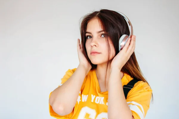 Adolescente aux cheveux longs foncés en t-shirt jaune écoutant de la musique ou baladodiffusion dans des écouteurs isolés sur fond gris — Photo