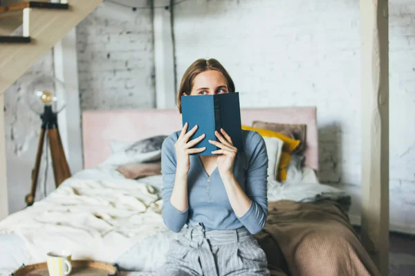 Junge Frau in der Nähe Gesicht Buch sitzt auf dem Bett im Loft Kunstwerk Atelierraum — Stockfoto